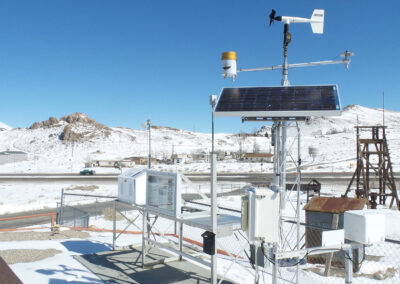 A weather monitoring station with solar panels, sensors, and a wind vane is set up in a snowy, mountainous area near a fenced property.
