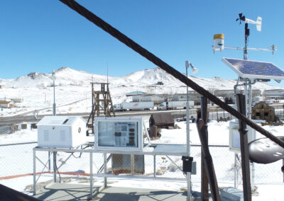 Weather monitoring equipment with solar panels and sensors is set up in a snowy, fenced area with mountains and buildings in the background under a clear blue sky.