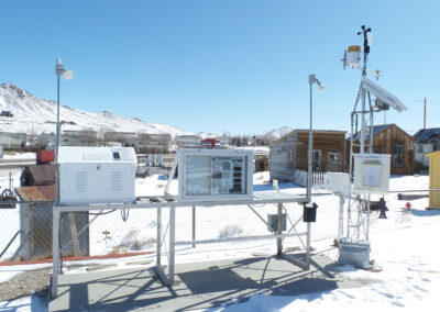 A weather monitoring station with various instruments and sensors is set up outdoors in a snowy area, with buildings and mountains in the background.