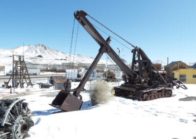 A large tracked excavator with a raised boom and bucket sits on snow-covered ground in an outdoor display, with buildings and mountains in the background.