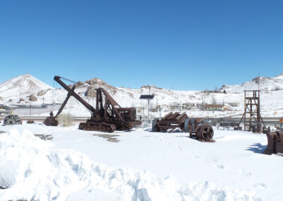 Rusty mining equipment sits on snow-covered ground with mountains and buildings in the background under a clear blue sky.