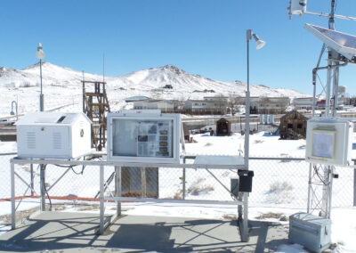 Outdoor weather monitoring station with sensors, solar panels, and display equipment, set against a snowy landscape with mountains and buildings in the background.