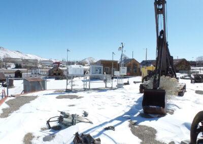 Snow-covered outdoor display with industrial equipment, including a large excavator bucket, surrounded by fencing and small buildings in the background under a clear blue sky.