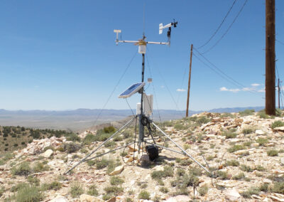 A weather station with solar panel and sensors stands on rocky terrain, surrounded by utility poles and desert landscape under a clear blue sky.