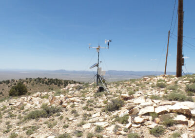 A weather monitoring station with instruments and a solar panel stands on a rocky hillside under a clear blue sky, overlooking a vast, arid landscape.
