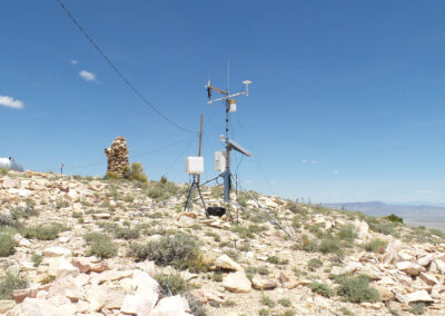 A weather monitoring station with various sensors and solar panels stands on a rocky hilltop under a clear blue sky.