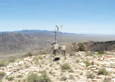 A weather station with instruments and sensors is set up on a rocky hilltop, overlooking a mountainous desert landscape under a clear sky.