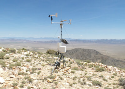A weather monitoring station stands on a rocky hilltop, equipped with sensors and instruments, overlooking a vast, arid landscape with mountains in the distance under a clear sky.